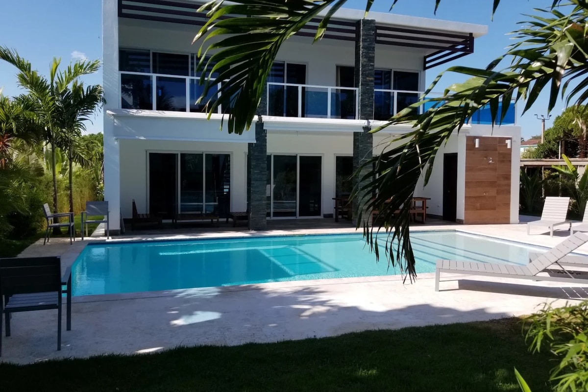 The villa presents a modern two-story structure surrounded by lush greenery. A large swimming pool is situated in the foreground, with sun loungers arranged nearby. Expansive glass doors lead to the terrace, enhancing the connection to the outdoor space.