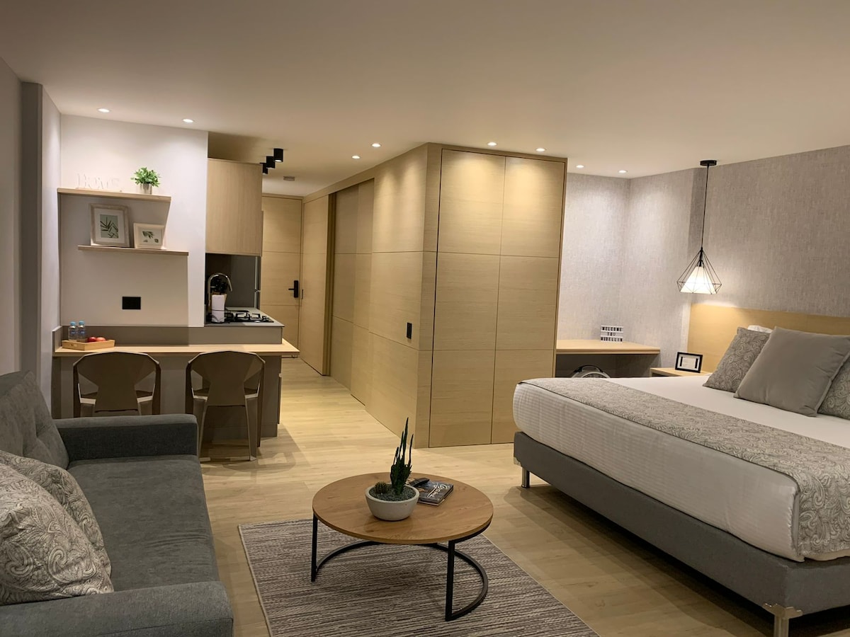 A well-furnished studio apartment features a spacious bed, a clean seating area with a grey sofa, and a round wooden coffee table. A kitchenette is present against the wall, along with a dining area equipped with two chairs. Soft lighting enhances the modern design.