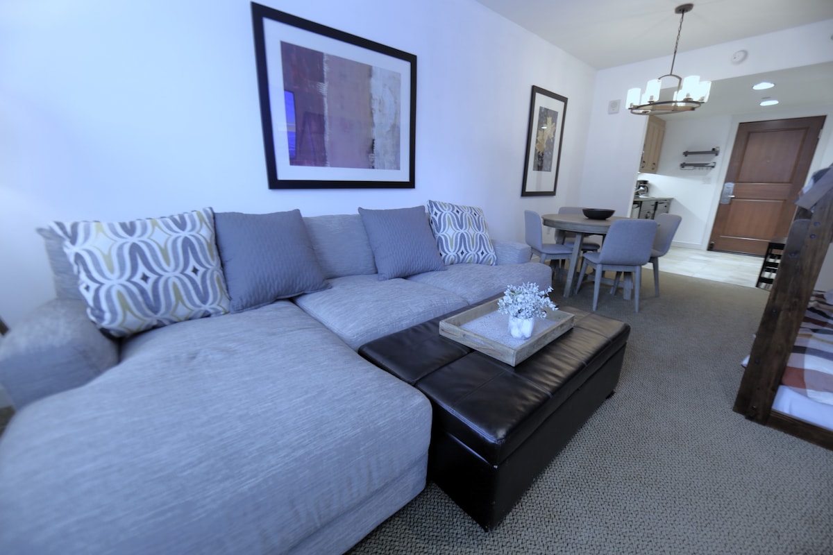 A spacious living area is showcased, featuring a large sectional sofa with a combination of light and dark upholstery. A wooden coffee table sits in front, displaying a decorative arrangement. In the background, a dining area is visible with a round table and four chairs.