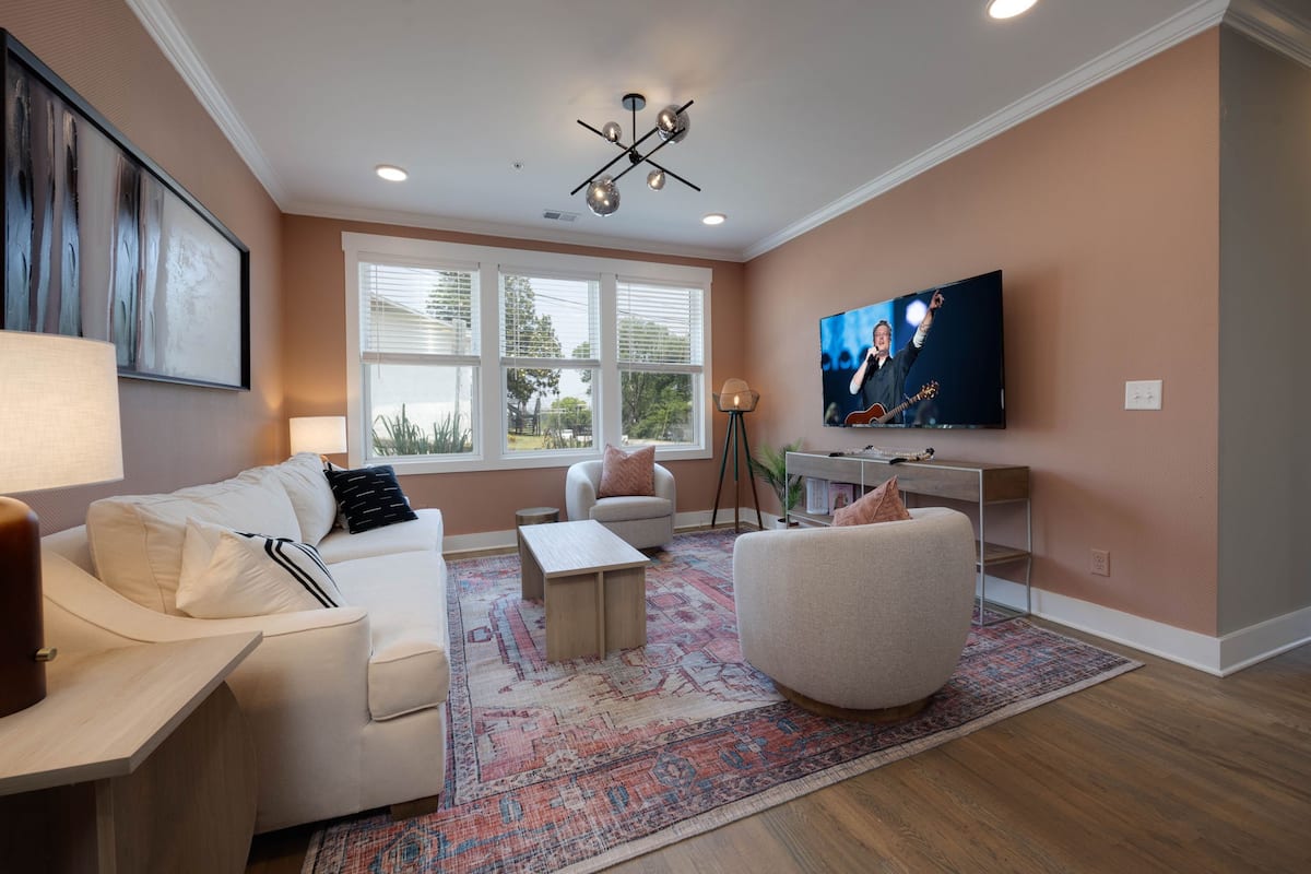 The living area features a cozy white sofa and two light-colored chairs arranged around a rustic coffee table. A large flat-screen TV is mounted on the wall, while natural light floods in through multiple windows adorned with minimalist blinds. A warm area rug adds a touch of comfort.