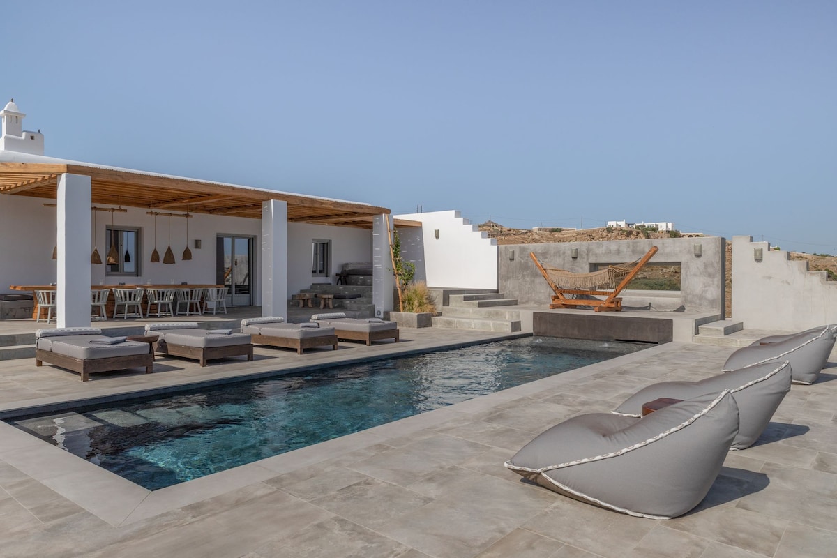 An inviting outdoor space features a serene swimming pool surrounded by cushioned loungers. A spacious terrace is visible, adorned with wooden furniture and hanging light fixtures. The area is bathed in natural light, highlighting the clean lines of the villa's modern architecture.