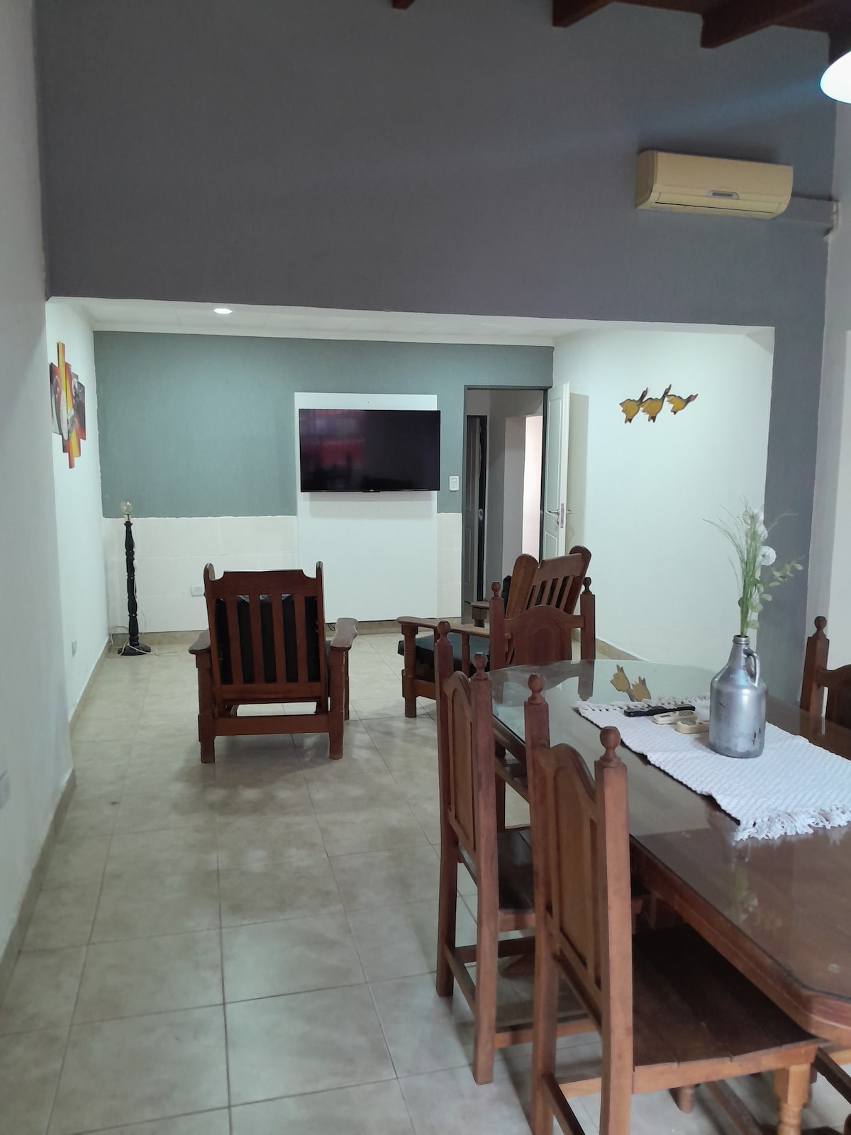 A spacious living area features wooden furniture, including a dining table with several chairs and comfortable seating options. A wall-mounted TV is visible, with light streaming through, enhancing the open layout. Decorative art pieces add a touch of color to the neutral-toned walls.