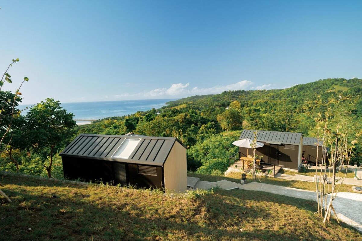 The image captures a scenic view of modern cabins nestled on a hillside, surrounded by lush greenery. The sparkling sea is visible in the distance, with a clear blue sky above. A pathway leads to the cabins, enhancing the connection with nature.