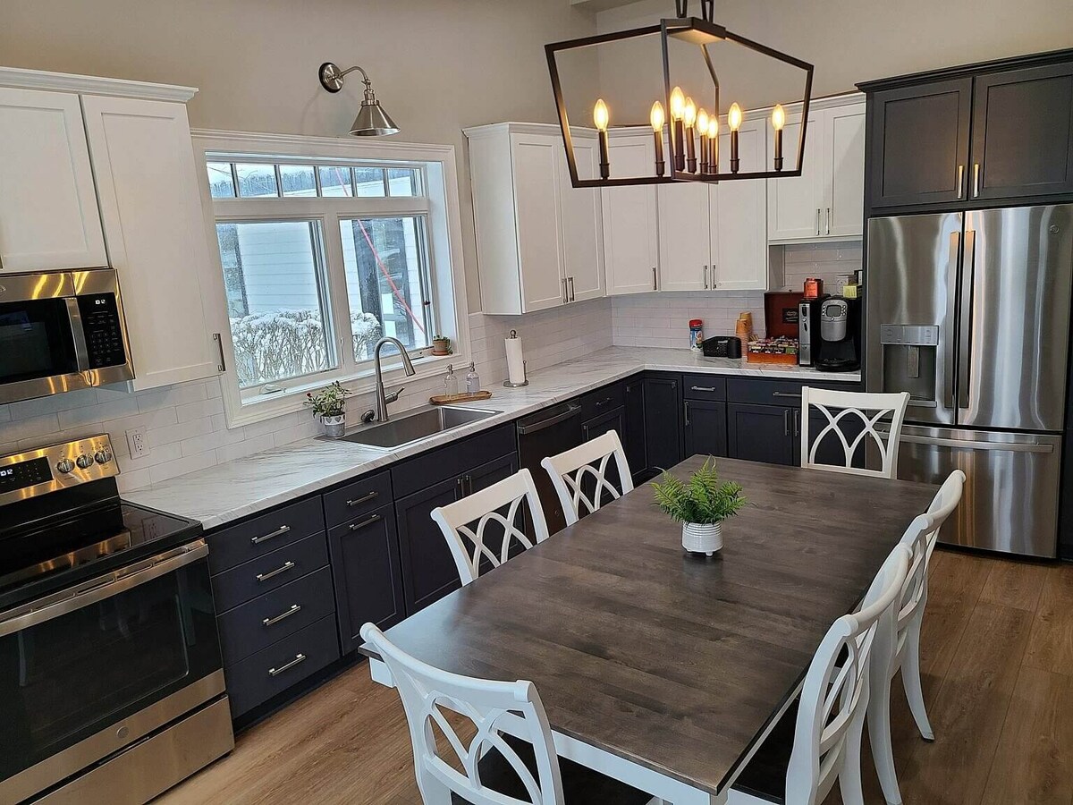 A modern kitchen is equipped with stainless steel appliances and a spacious dining table surrounded by white chairs. A large window allows natural light to illuminate the area, and a decorative plant centerpieces the table, enhancing the welcoming atmosphere.