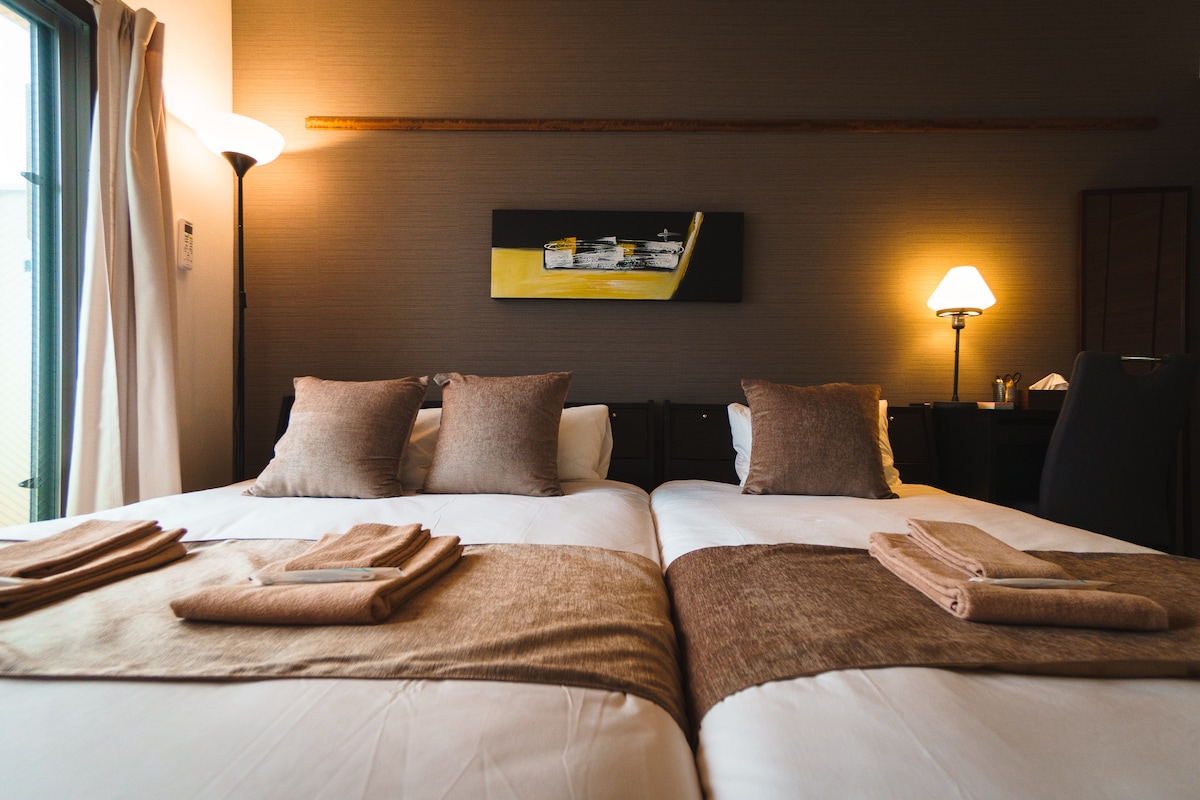 A well-appointed sleeping area is displayed with two spacious beds, each adorned with neutral-toned linens and decorative pillows. Natural light filters through a nearby window, illuminating a simple artwork above the beds. A lamp softly brightens the space, contributing to a calming ambiance.