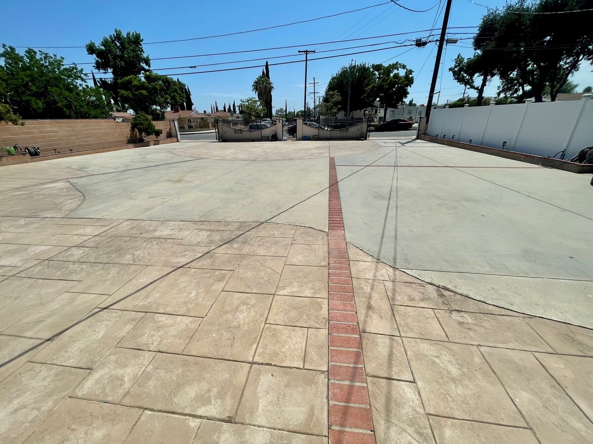 A spacious outdoor area is shown, featuring a large concrete patio with a decorative brick pathway running through it. The surrounding spaces include greenery and fencing, providing a sense of privacy and openness.