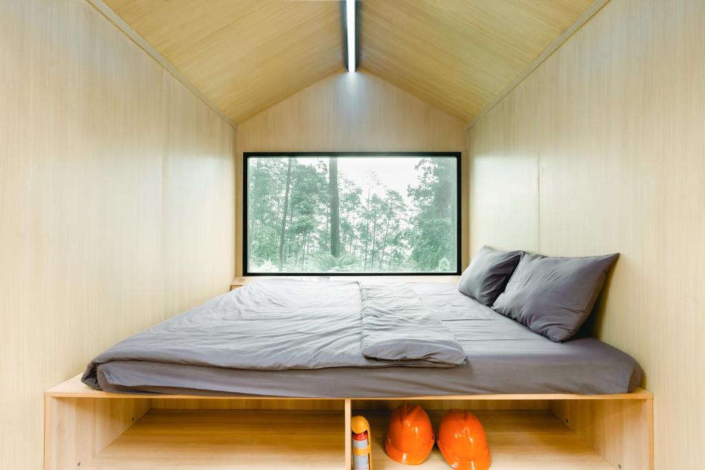The cabin interior features a spacious bed covered with grey bedding. Below the bed, orange safety helmets and a canister are visible. A large smart glass window provides a view of the surrounding forest, while wooden walls and ceiling create a natural ambiance.