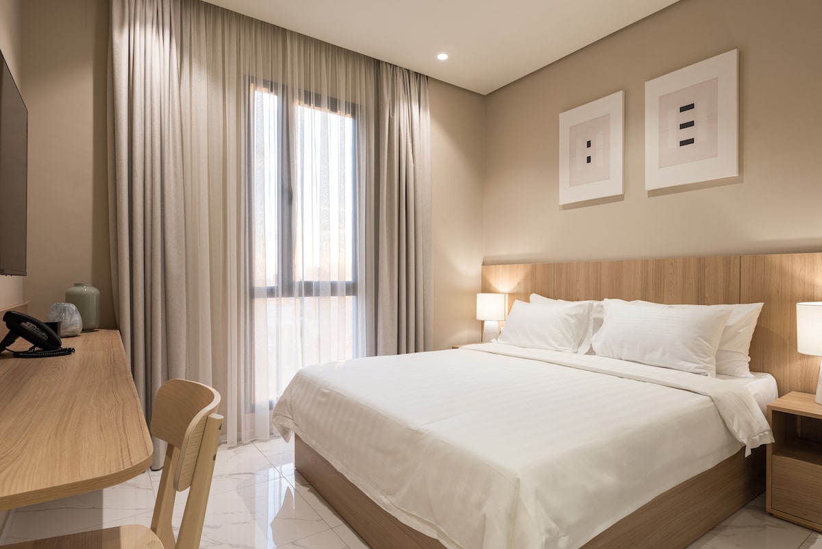 A contemporary bedroom is showcased, featuring a spacious bed with crisp white linens. Light wood furniture includes a desk and bedside tables, each with soft-lit lamps. Sheer curtains allow natural light to filter in through the window, highlighting the minimalist artwork on the walls.