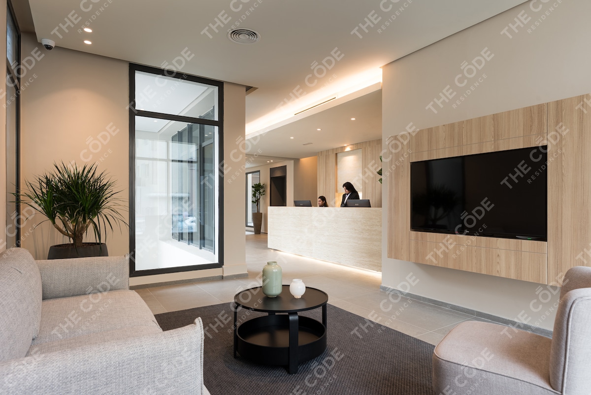 The lobby area features contemporary seating arrangements with soft gray sofas facing a round black table. A large flat-screen television is mounted on the light wood wall, while natural light floods in through a large glass door, which opens to the exterior.