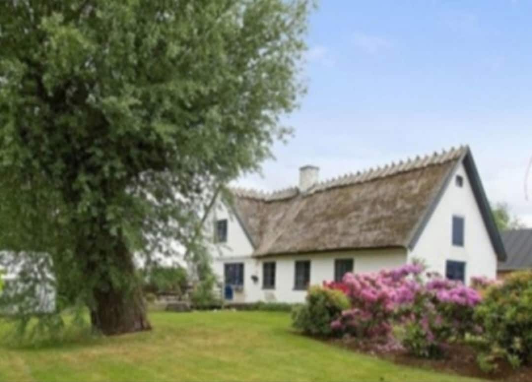 A historic farmhouse from 1867 is surrounded by lush greenery and colorful flowering shrubs. The thatched roof and white exterior are complemented by ample outdoor space, inviting a sense of tranquility. A large tree provides shade, enhancing the outdoor setting.