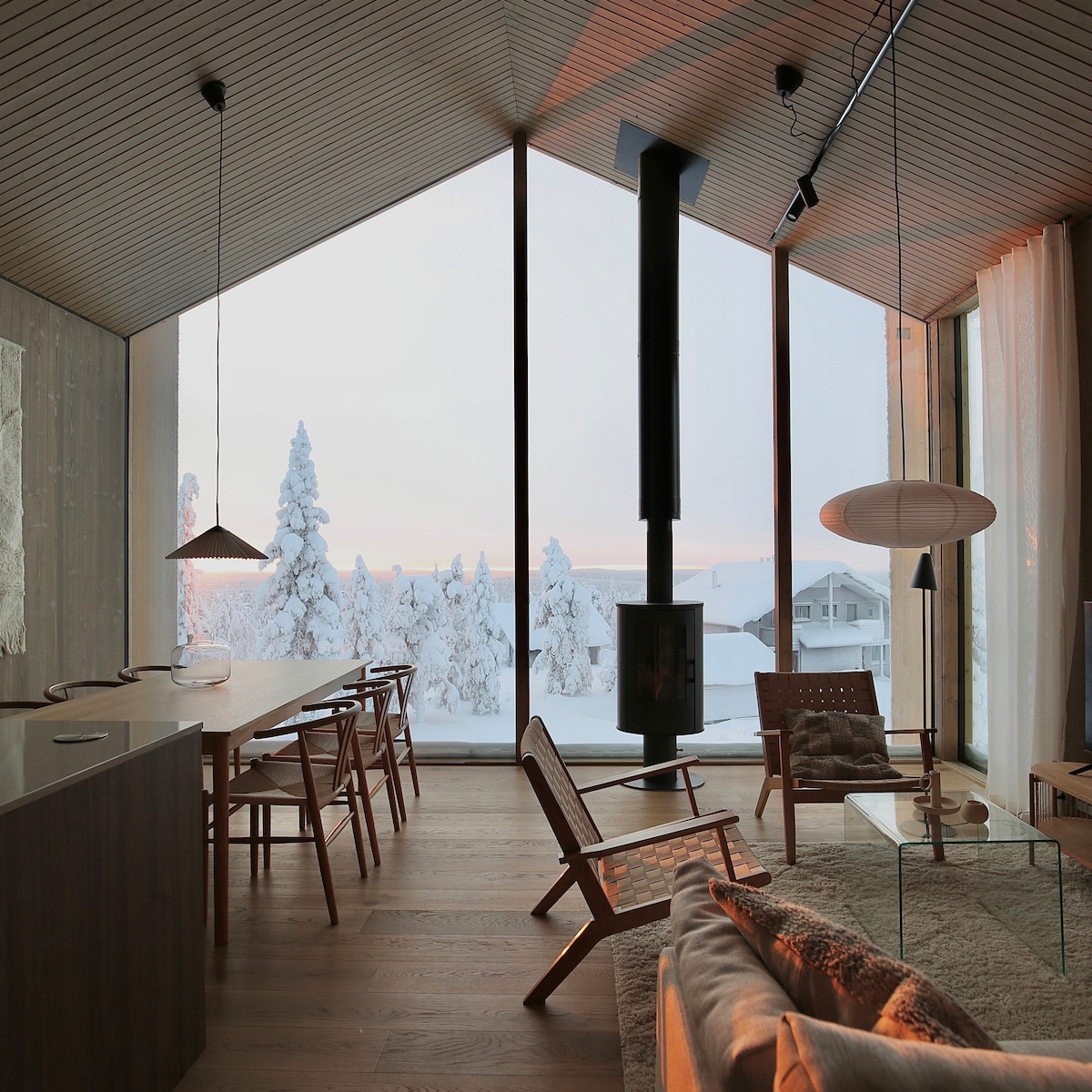 A modern living area features large panoramic windows that reveal a snowy landscape. A sleek black fireplace serves as a focal point, with seating arranged thoughtfully around a glass table. Wooden dining chairs are positioned near a long table, providing a harmonious space for gatherings.