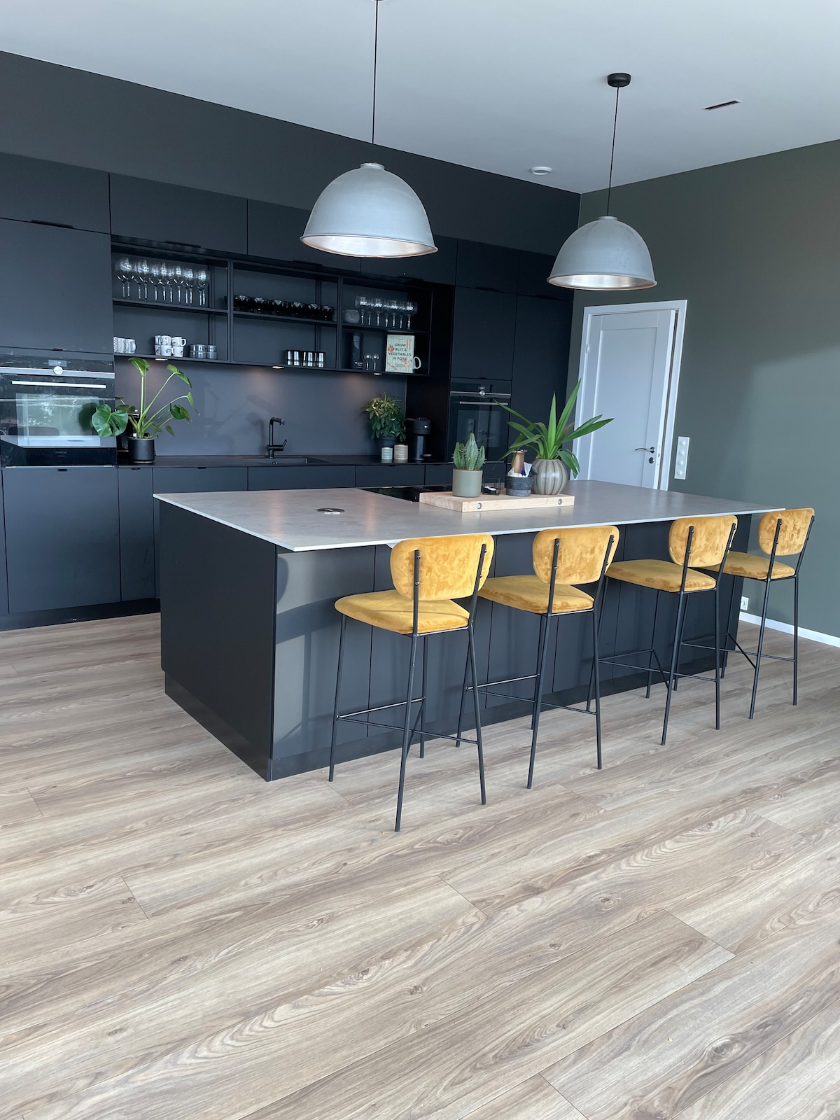 The kitchen features a modern island with a light countertop and four yellow barstools. Dark cabinetry and built-in appliances create a sleek look, while plants add a touch of greenery. Pendant lights hang above the island, illuminating the space.
