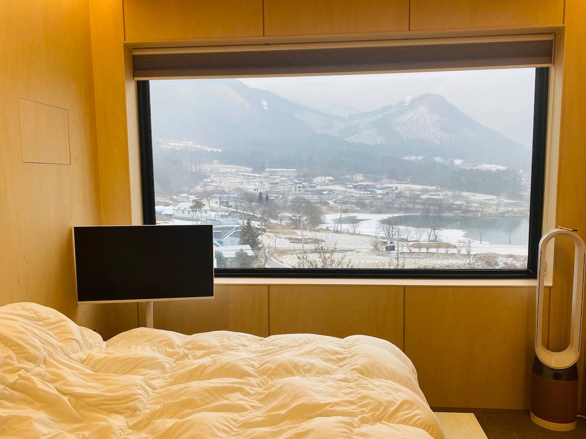 A spacious sleeping area features a large window offering views of snow-covered mountains and valleys. A soft, crumpled duvet lies atop the bed, while a sleek television is positioned nearby, creating a comfortable ambiance. Natural light fills the room, enhancing the serene atmosphere.