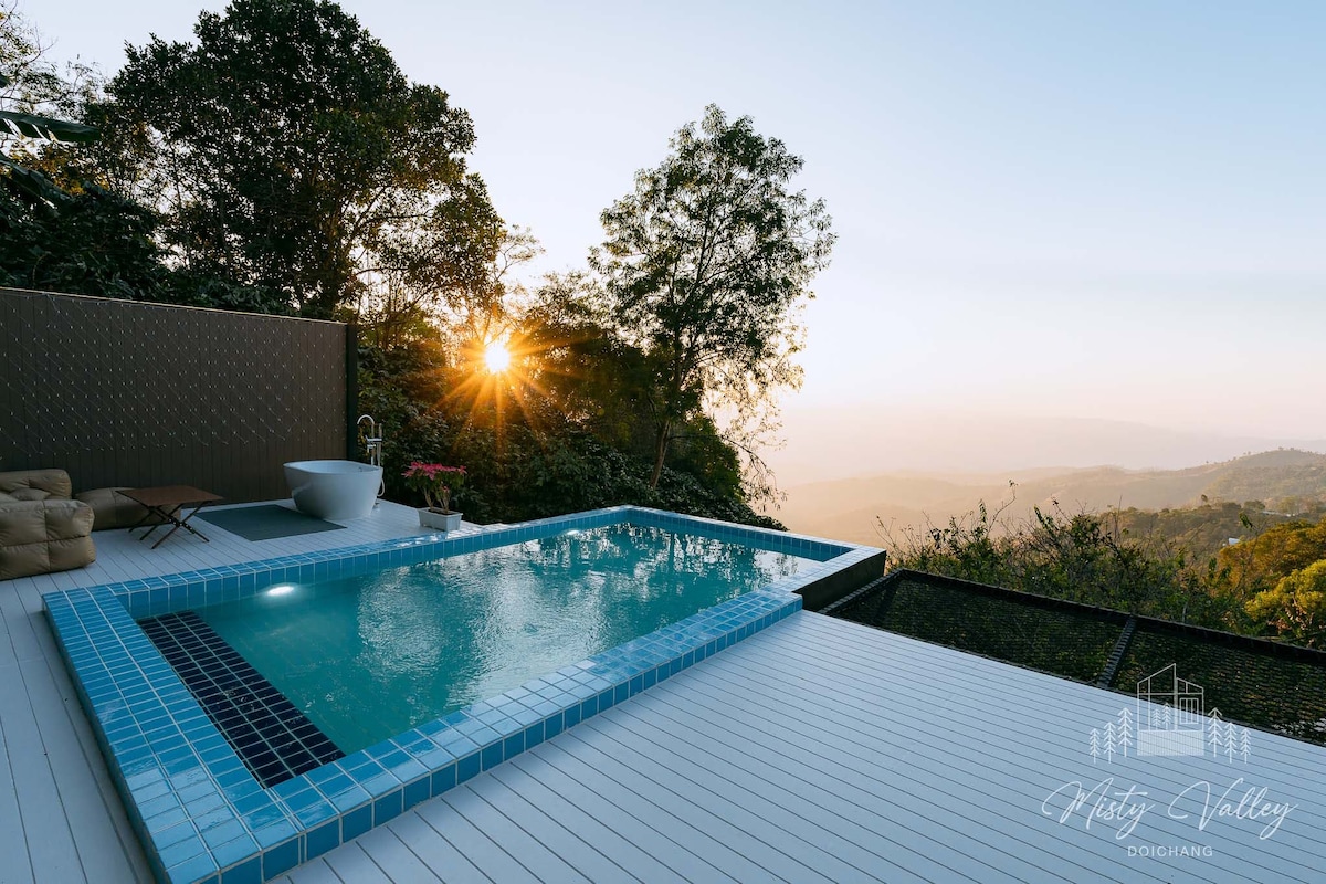 A private pool is positioned on a deck with scenic mountain views, featuring a sunken bathtub nearby. The soft glow of sunset illuminates the surroundings, while lush greenery frames the area, creating a serene outdoor retreat.