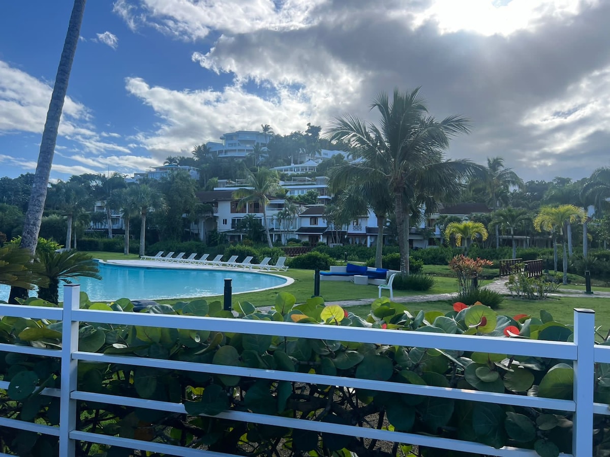 A spacious pool area is surrounded by lush greenery and palm trees, with lounge chairs positioned along the edge. In the background, multi-story buildings are seen, nestled among vibrant tropical landscaping under a partly cloudy sky.