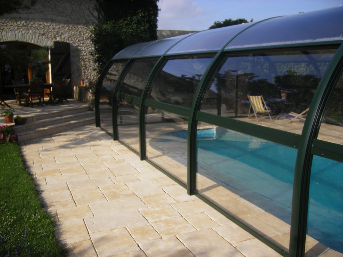 A covered swimming pool is visible, surrounded by a stone patio with neatly arranged tiles. The pool is enclosed by a transparent structure, allowing for ample natural light. Comfortable outdoor seating is positioned nearby, offering a relaxing space in the lush garden setting.