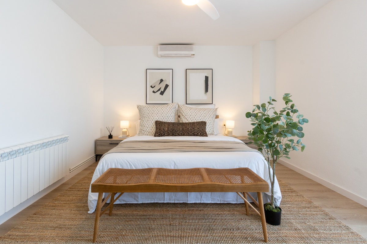 A spacious bedroom features a large bed with neutral-toned bedding and accent pillows. Two framed artworks adorn the white walls, complemented by bedside lamps that provide soft lighting. A wooden bench is positioned at the foot of the bed, with a potted plant adding a touch of greenery.