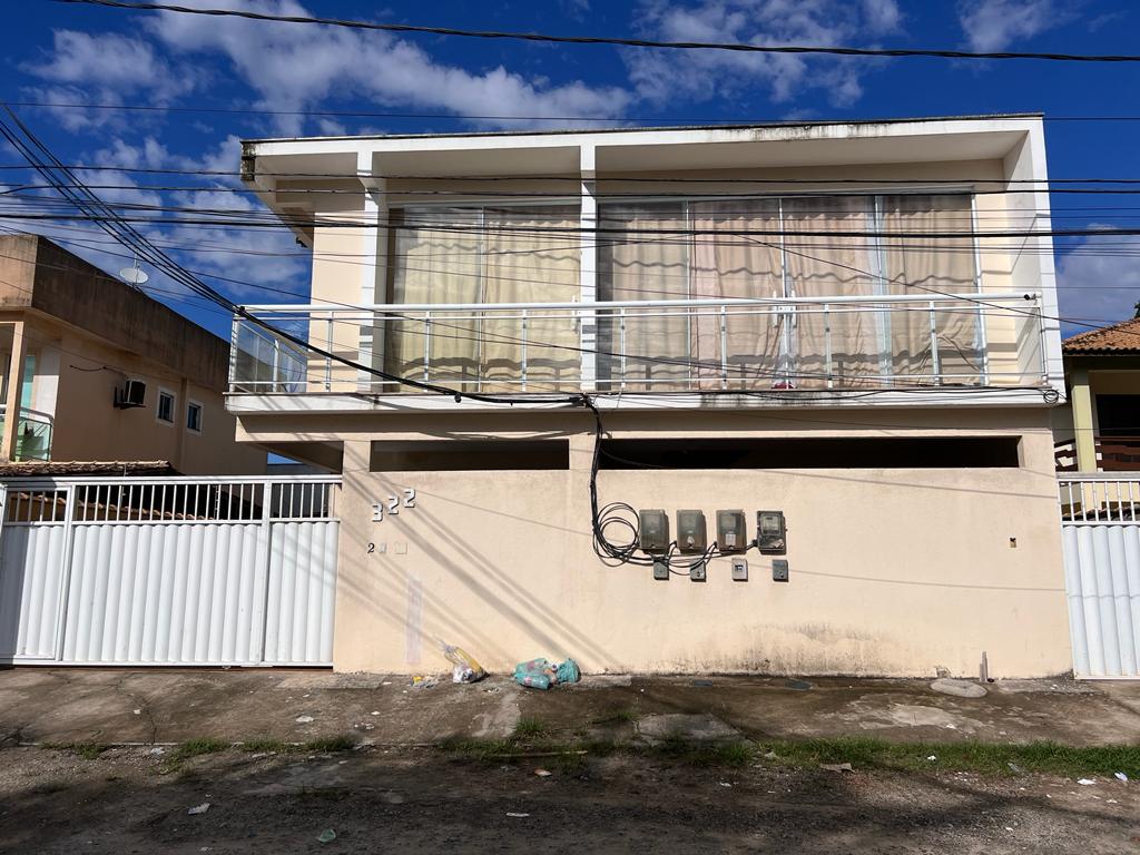 A modern two-story building is shown with a glass balcony on the upper floor, allowing ample natural light into the interior. The façade features neutral tones and utility meters are mounted on the wall. A clean white fence surrounds the property.