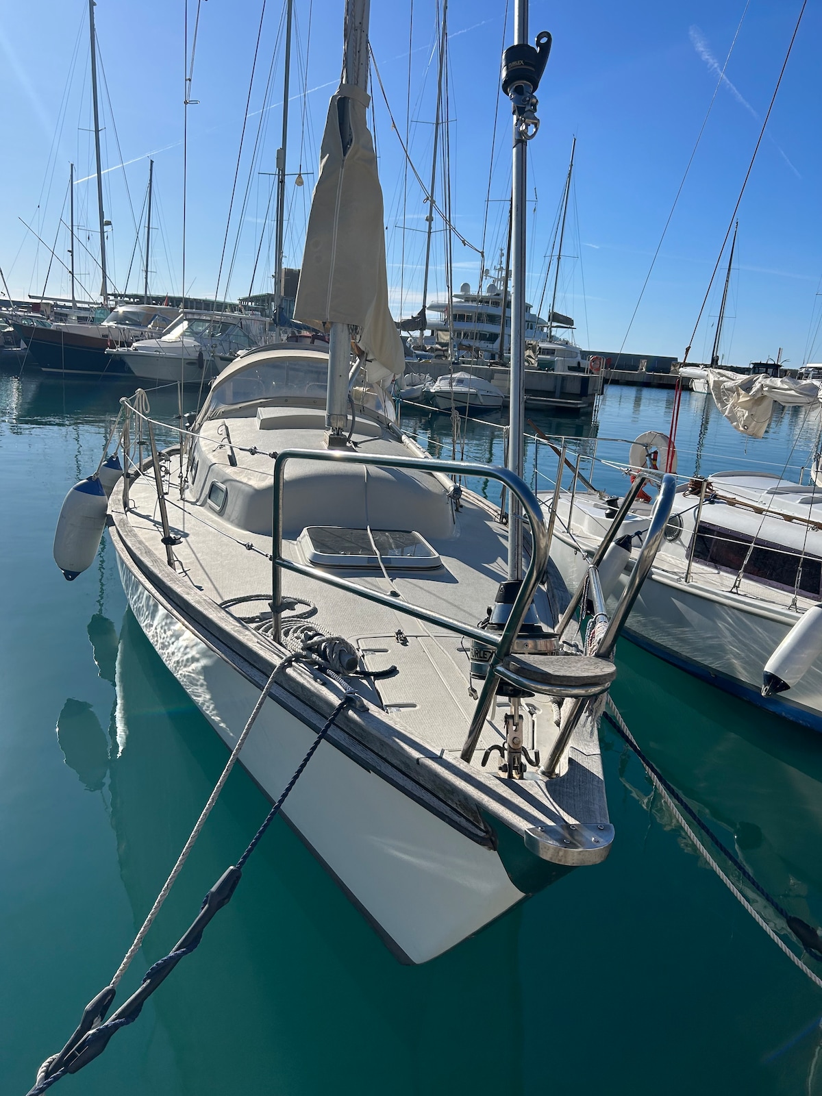 A sailboat is docked in a modern marina, its white hull reflecting the sunlight on clear blue water. The equipment on the deck includes a winch and ropes, with masts and sails visible in the background, surrounded by other boats.