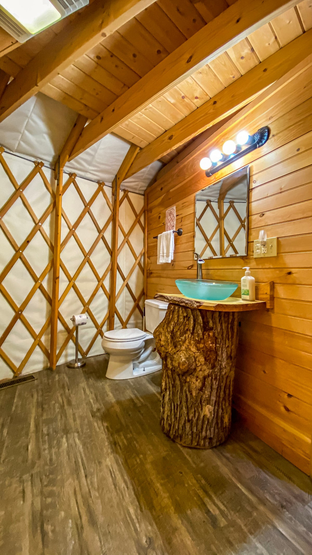 A bathroom within the yurt is highlighted by a wooden sink basin and a mirror mounted above it. Natural wood finishes adorn the walls, and a toilet is positioned nearby. Ample lighting is provided by a multi-bulb fixture overhead, enhancing the warm atmosphere.