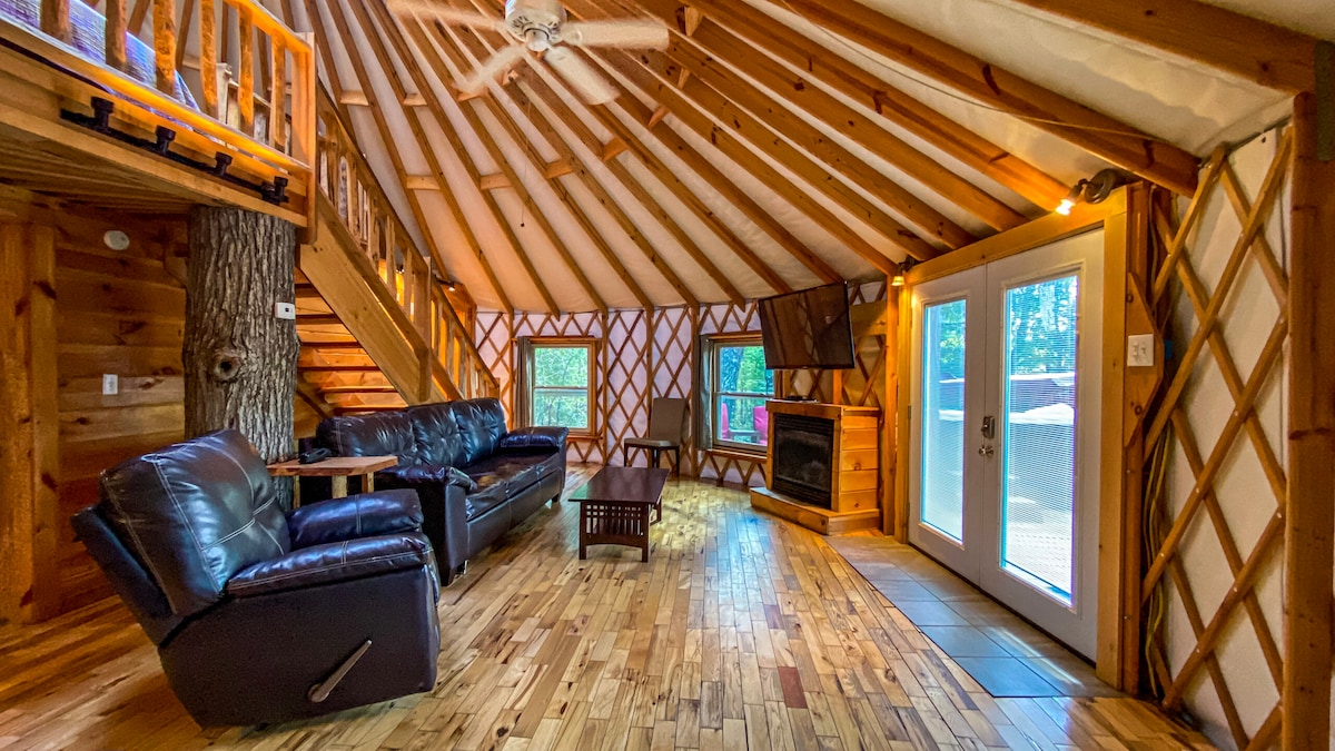The interior of the yurt features wooden beams and a vaulted ceiling, enhancing the spacious atmosphere. Comfortable leather seating is arranged around a central coffee table, with a flat-screen TV mounted nearby. Natural light floods in through glass doors leading to the outdoor deck.