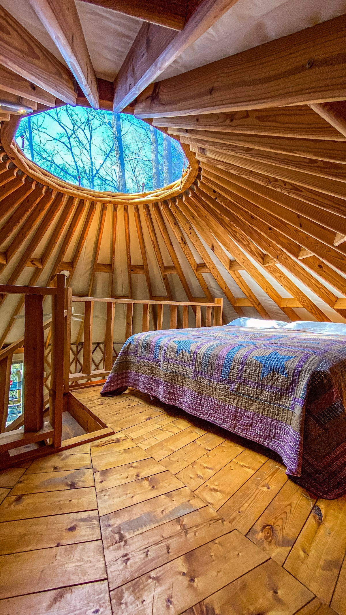 A cozy loft space is highlighted by natural wood beams and a circular skylight that allows light to illuminate the area. A queen-sized bed, adorned with a textured blanket, sits on a wooden floor, enhancing the serene atmosphere surrounded by trees.