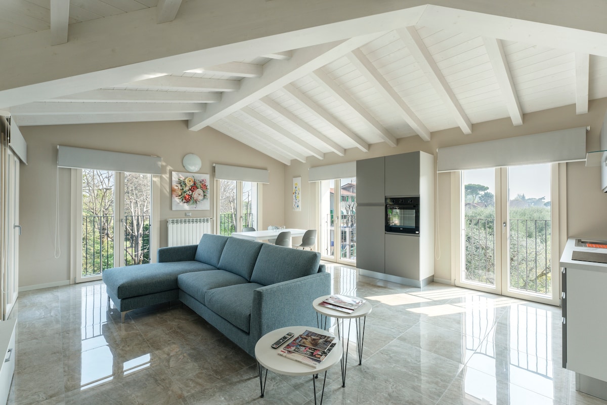 A spacious living area features high, exposed beam ceilings and large windows that allow natural light to fill the room. A modern sectional sofa and a coffee table are positioned on polished tile flooring. A dining table is visible in the background, creating an inviting atmosphere.