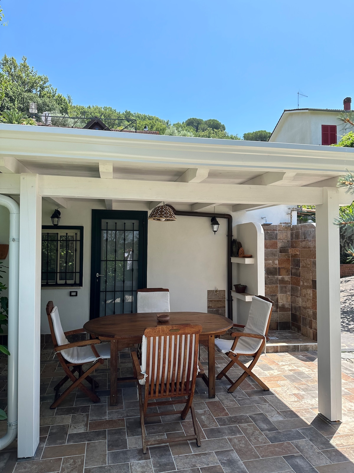 An outdoor patio area features a round wooden table surrounded by four light-colored chairs. A decorative lamp hangs from the ceiling above the table, and stone tiles cover the ground. Lush greenery is visible in the background, creating a serene environment.
