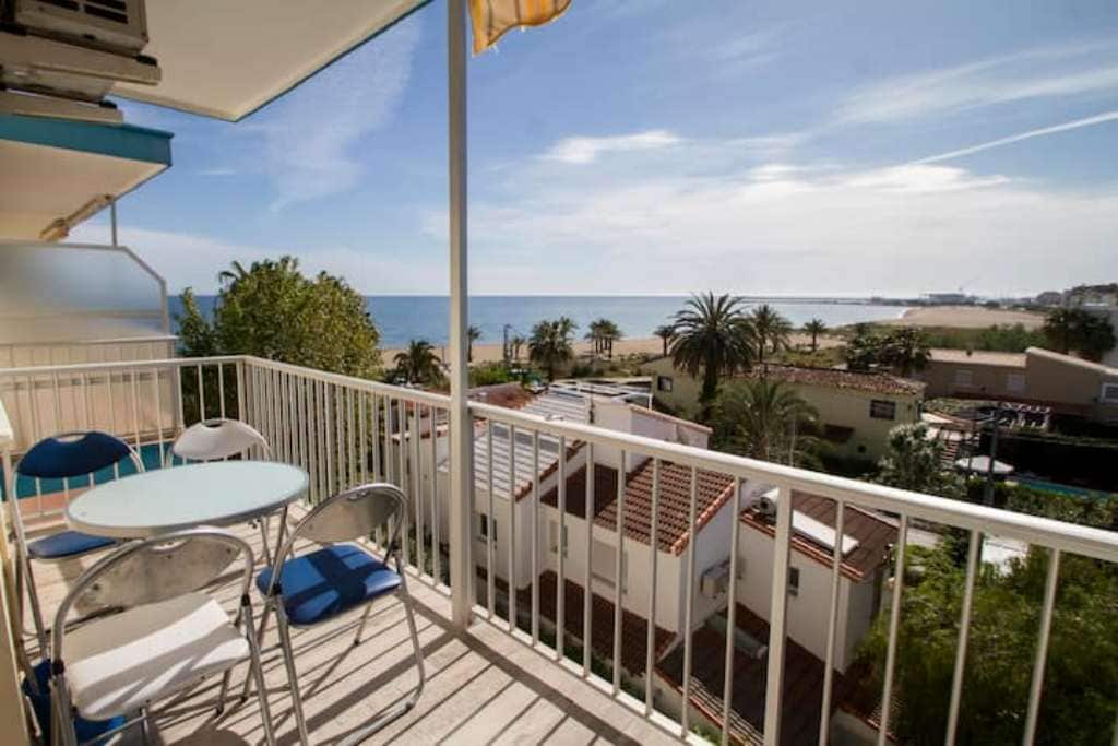 A balcony is visible overlooking the beach and ocean, featuring a small round table and four metal chairs. Palm trees are seen below, with rooftops and the shoreline in the background, providing a serene coastal view.