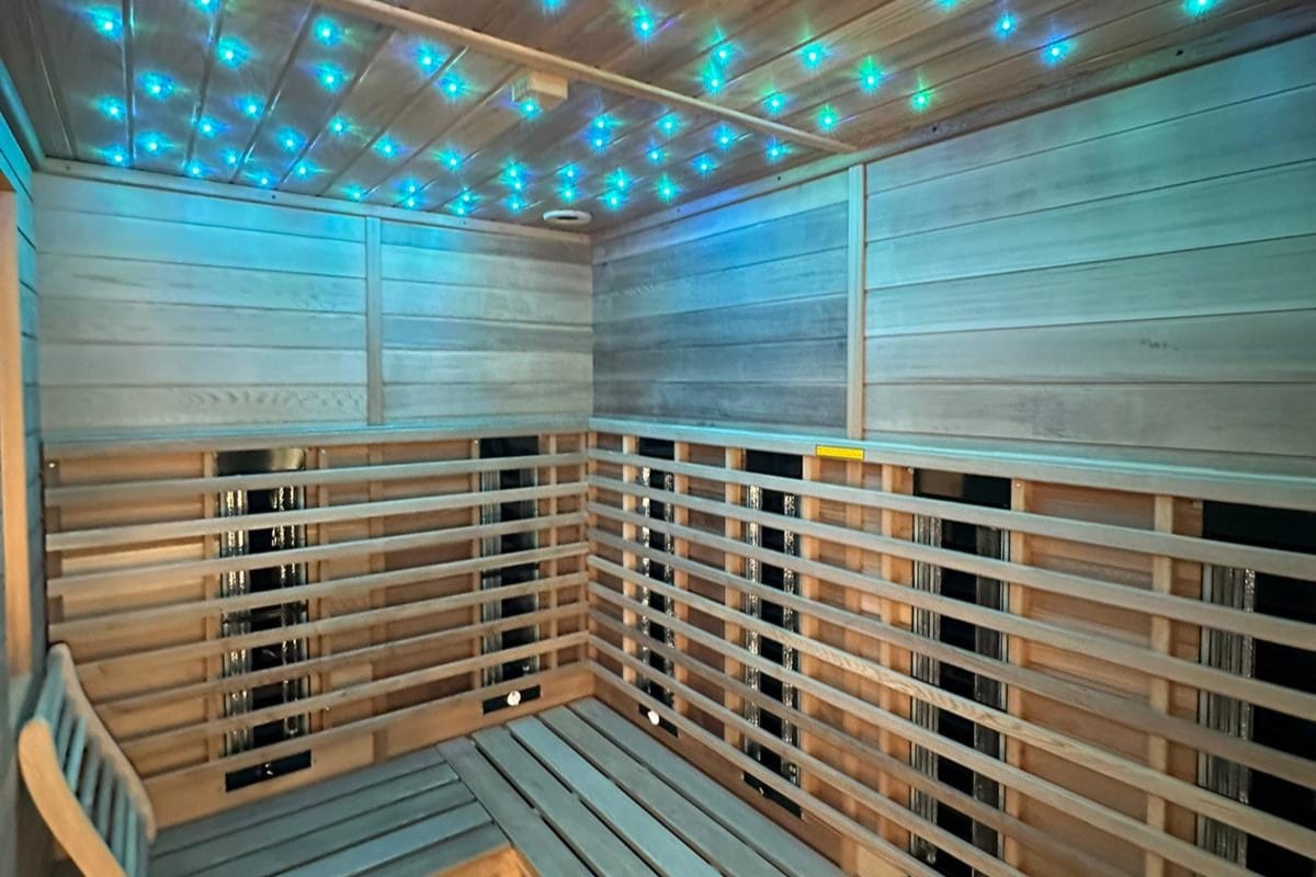 The sauna features wooden slatted walls and a bench, illuminated by soft blue and green lights on the ceiling. The calming environment is designed for relaxation, with ample space for seated guests.