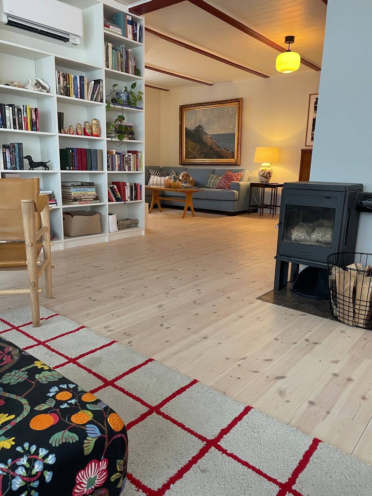A cozy living area is visible, featuring a mix of bookshelves and comfortable seating. A sofa is positioned across from a small wood-burning stove, while a patterned rug adds a touch of warmth. Natural light enhances the inviting space.
