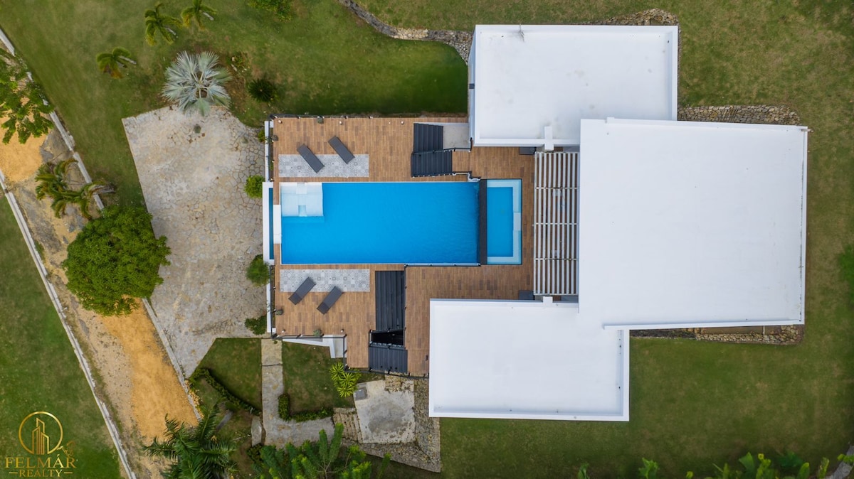 An aerial view of a modern villa showcasing a long rectangular pool bordered by wooden decking and multiple lounge chairs. Lush greenery surrounds the property, with pathways leading to different areas. The structure features a clean, white exterior and a spacious layout.