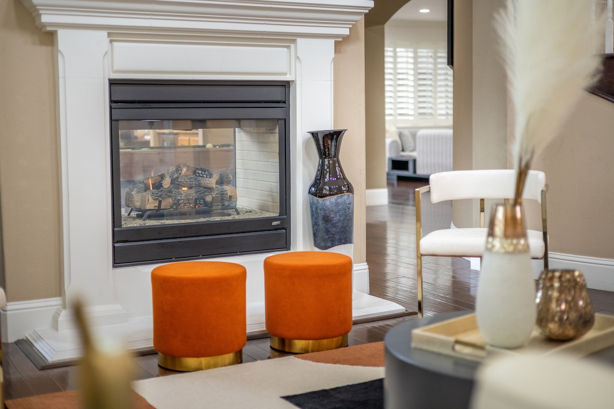 A modern living area features a sleek fireplace, framed by a clean white mantel. Two vibrant orange stools with gold bases sit in front, complementing a neutral rug. A decorative vase and various accents are arranged on a coffee table, adding an inviting touch to the space.