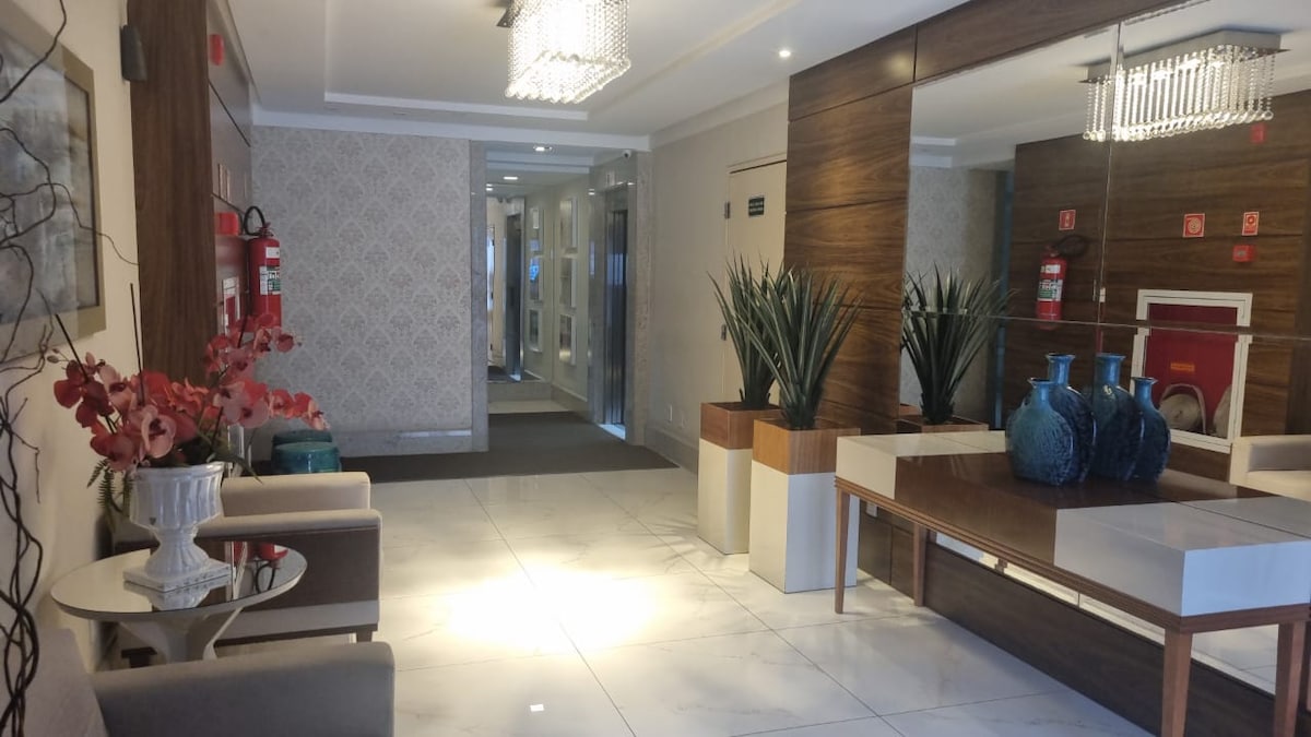A spacious lobby area features marble flooring and wooden accents. Modern furnishings include two armchairs and a large table topped with decorative vases and greenery. The area is illuminated by a chandelier above, while mirrored walls create an airy feel.