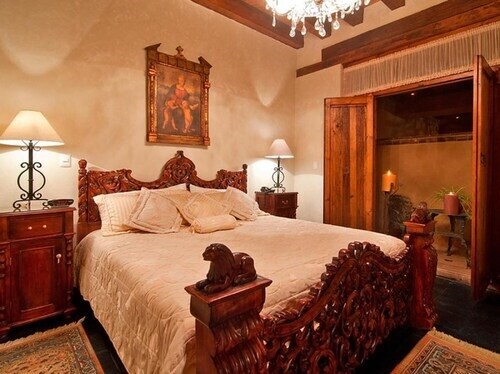 A spacious bedroom features a large wooden king-size bed adorned with intricate carvings. Two bedside lamps provide soft lighting, and a framed artwork hangs above the bed. Rich wooden finishes are complemented by a chandelier, creating an elegant atmosphere.