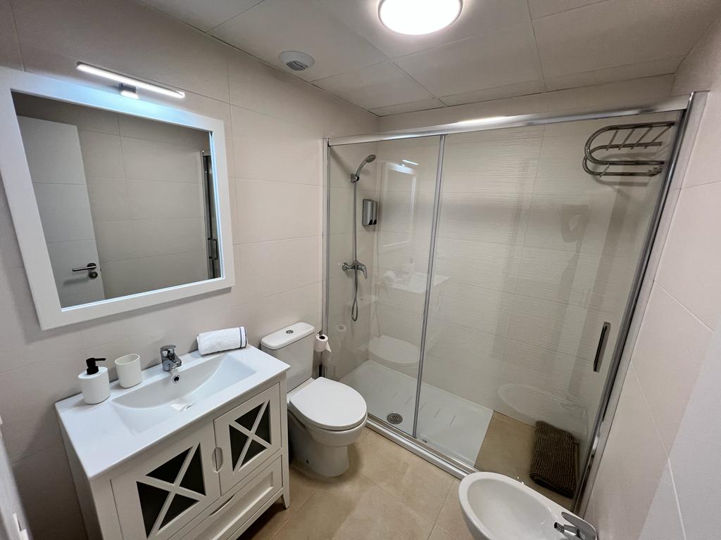 A modern bathroom features a glass shower enclosure with a rainfall showerhead. A white vanity with storage is positioned under a mirror, complemented by neutral tiled walls. Essential fixtures include a toilet, bidet, and a towel rack, creating a functional and tidy space.