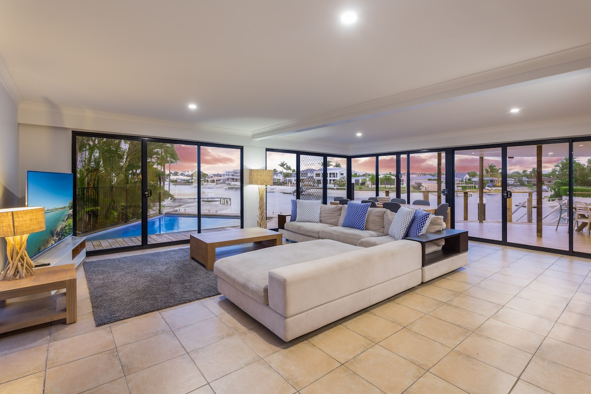 A spacious living area showcases large glass windows that provide views of the waterfront and sunset. A light-colored sectional sofa is complemented by decorative cushions, and a large area rug adds warmth to the tiled floor. A flat-screen TV is positioned on one wall.