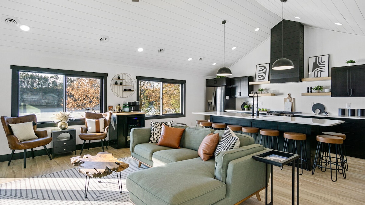 The open-concept living area features modern furnishings, including a green sectional sofa and two brown accent chairs. Large windows provide abundant natural light and views of the outdoors. A dining island with bar stools complements the gourmet kitchen, equipped with sleek black appliances.