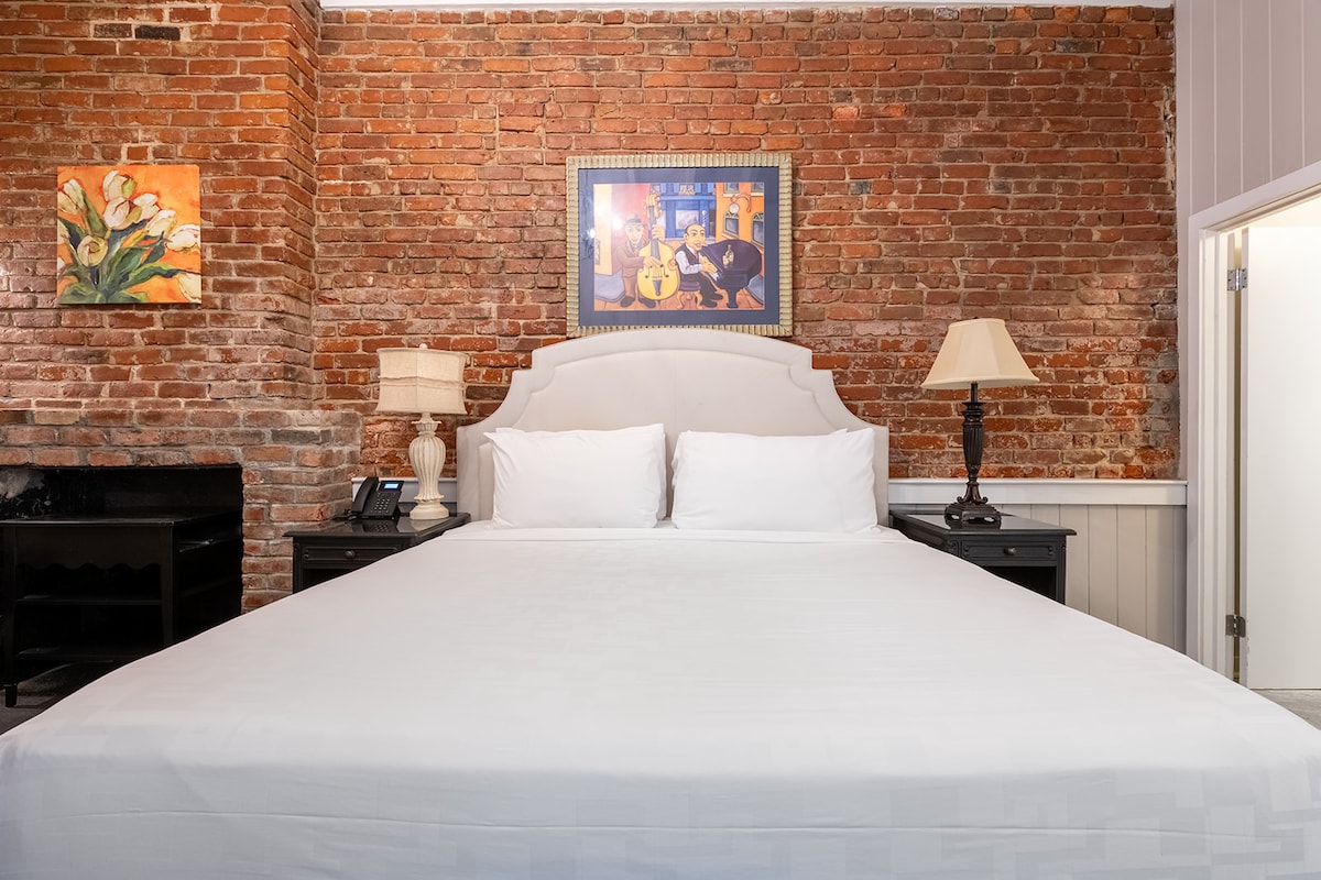 A spacious bed is centered against a textured brick wall, complemented by light bedding. A framed artwork is displayed above the bed, while side tables with lamps are positioned on either side. A doorway is visible in the background.