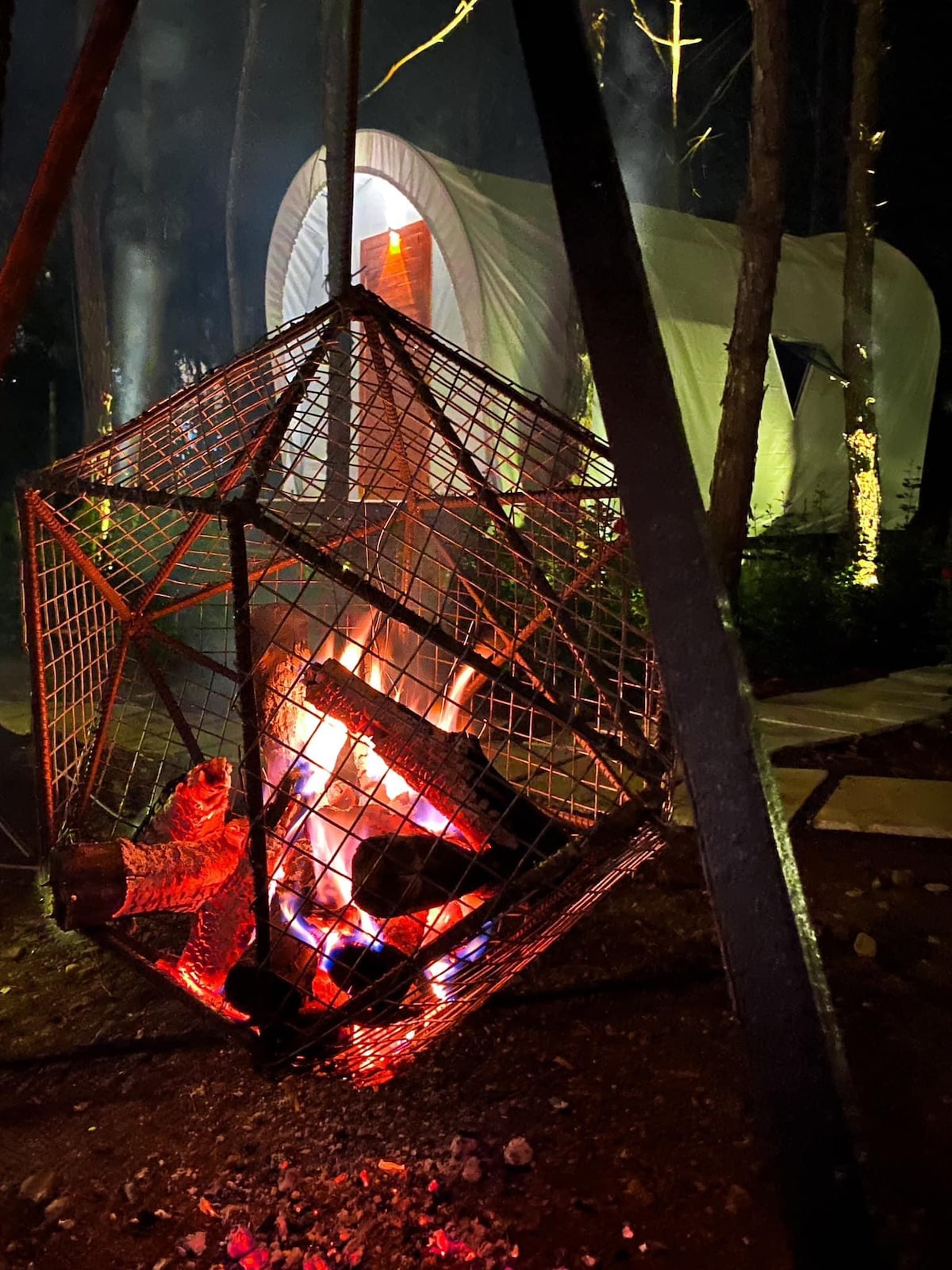 A suspended metal fire pit is seen, containing bright flames and glowing embers. In the background, a tent-like structure is softly illuminated, nestled among trees in a dark setting, enhancing the atmosphere of serenity.