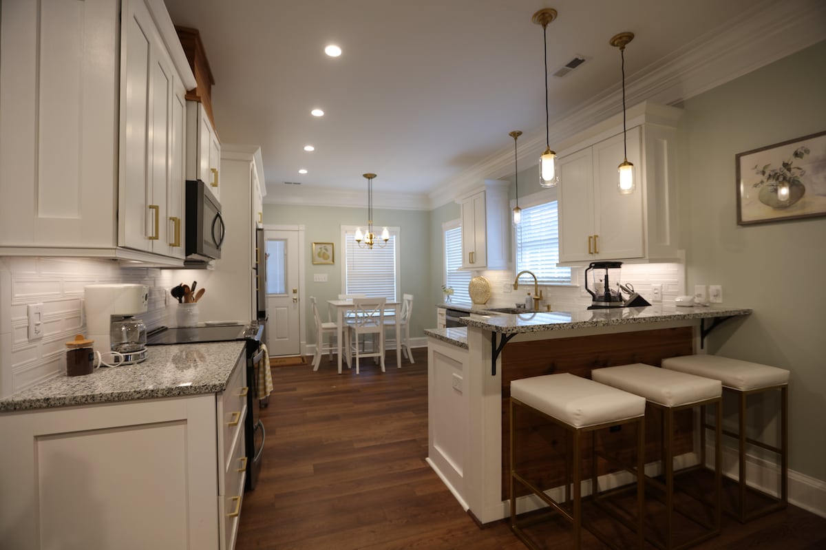 A modern kitchen is showcased with white cabinetry, granite countertops, and pendant lighting. Bar stools are positioned at a kitchen island. A dining area is visible in the background, featuring a small table and four chairs. Natural light fills the space through the windows.