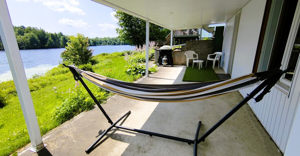 A hammock is set up on a covered patio, providing a relaxing space. Two white chairs are positioned nearby, with a grassy area leading to the tranquil waters of the lake. Lush greenery lines the shore, enhancing the peaceful outdoor setting.