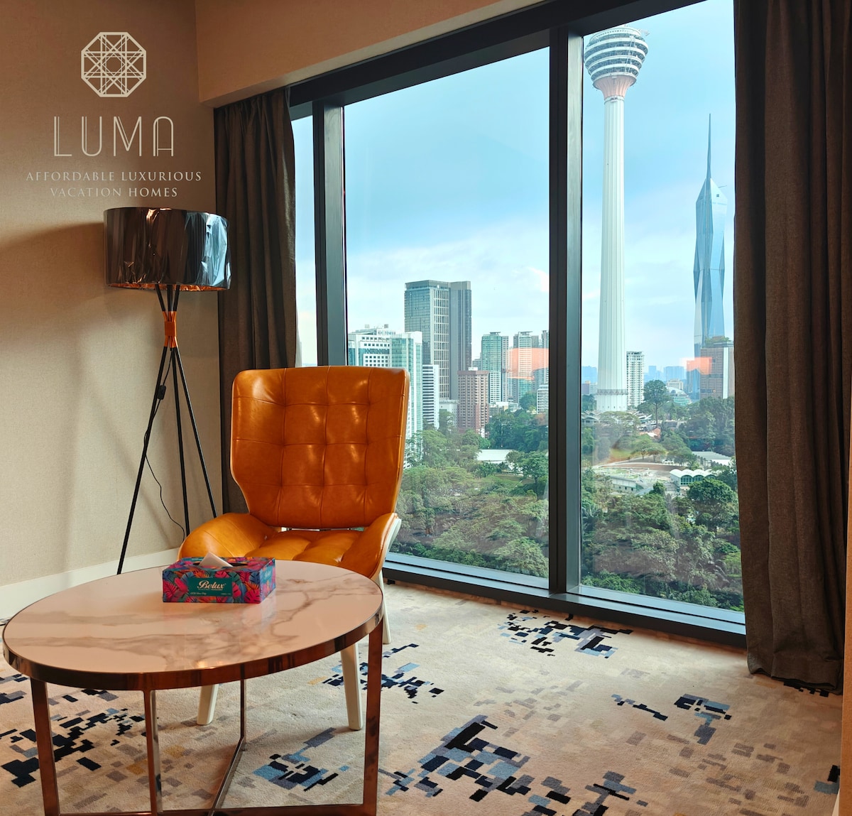 A modern seating area is presented with a stylish orange chair and a round table. Floor-to-ceiling windows reveal a view of the iconic KL Tower and city skyline, while natural light enhances the space.