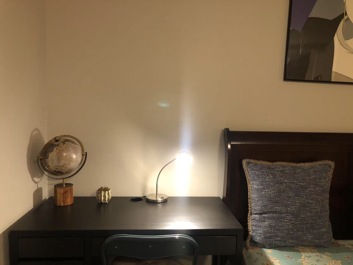 A simple desk is positioned adjacent to a spacious bed. A globe sits on the desk beside a modern lamp. A decorative pillow with a blue pattern rests on the bed, while a piece of artwork is visible on the wall above.