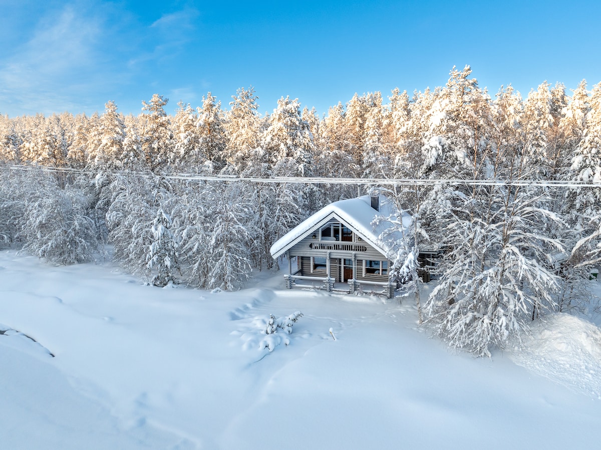 A winter scene captures a wooden cabin surrounded by a snowy landscape. The cabin features a sloped roof and a wide porch, while snow-covered trees provide a tranquil backdrop. The serene atmosphere is complemented by clear blue skies.