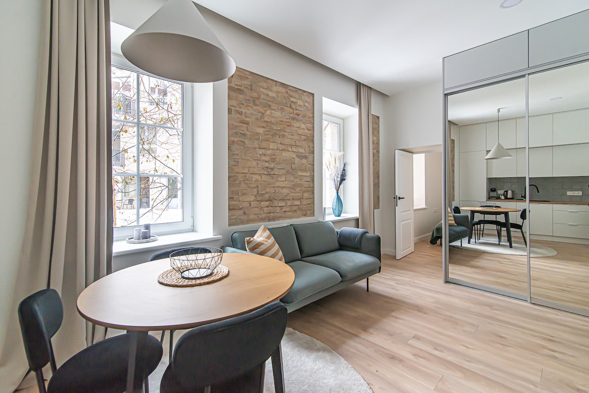A modern living area features a comfortable sofa and a round dining table with four chairs. Large windows allow natural light to fill the space, while a soft area rug complements the wood flooring. Decorative plants add a touch of elegance to the atmosphere.