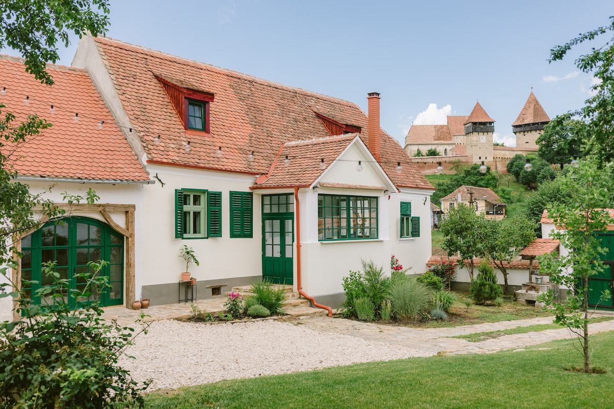 A traditional Saxon house features charming green shutters and a welcoming entrance. The rustic stone pathway leads to a garden area, with lush greenery surrounding the exterior. The historical fortified church stands prominently in the background, complementing the serene rural landscape.