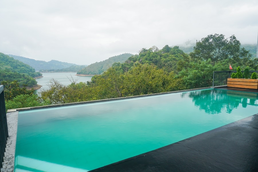 An infinity pool reflects the serene landscape, seamlessly blending with the surrounding mountains and lake. Lush greenery frames the scene, enhancing the tranquil atmosphere. Soft hues from the cloudy sky are mirrored in the water's surface, creating a calm and peaceful setting.