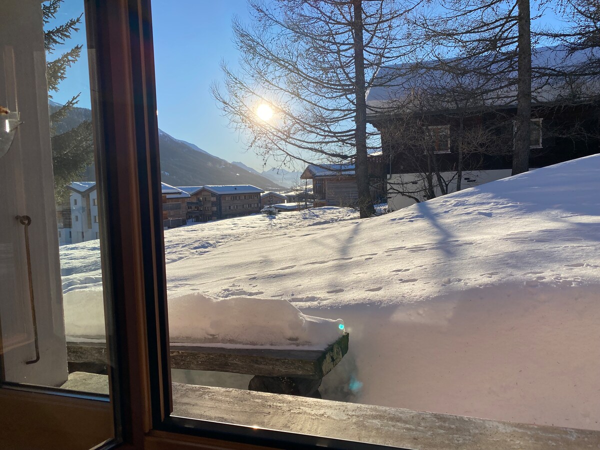 A view from the window showcases a snow-covered landscape bathed in sunlight. The distant mountains are visible, with trees casting long shadows on the ground. The scene captures the serenity of a winter day and a bench nestled in the snow.