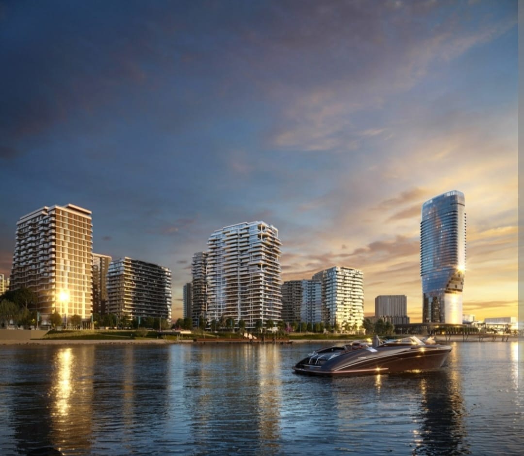 The image captures a tranquil riverside view featuring modern buildings reflecting the sunset. A sleek boat is seen cruising on the water, with soft golden light illuminating the scene and enhancing the serene atmosphere of the developing urban landscape.